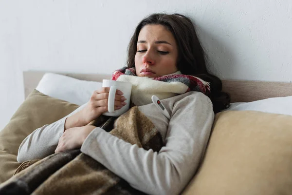 Хвора жінка лежить під картатою ковдрою з чашкою чаю — стокове фото