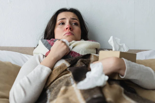 Upset woman lying under plaid blanket near crumpled paper napkins, blurred foreground — Stock Photo