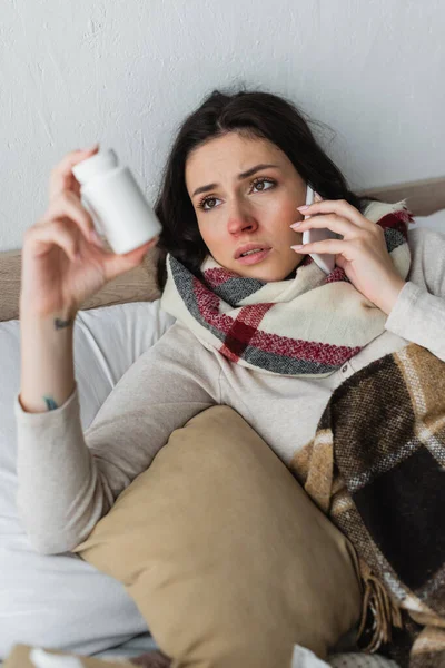 Mulher doente com pílulas recipiente falando no telefone celular enquanto deitado sob manta xadrez — Fotografia de Stock