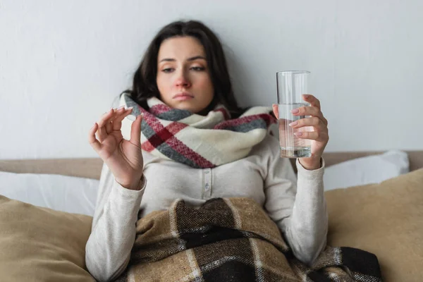 Хвора і сумна жінка лежить у ліжку зі склянкою води і дивиться на таблетки — стокове фото