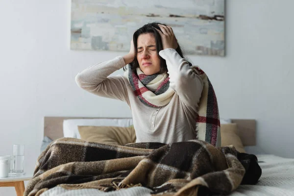 Kranke Frau in warmem Schal berührt Kopf, während sie unter Kopfschmerzen im Schlafzimmer leidet — Stockfoto