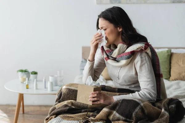 Sick woman in warm scarf sitting under plaid blanket and sneezing in paper napkin — Stock Photo