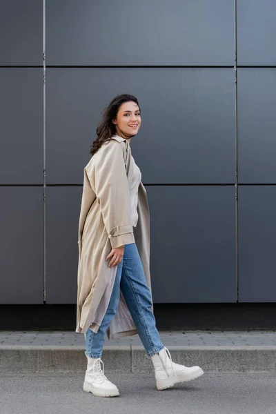 Jeune femme en manteau long et bottes en cuir blanc regardant la caméra près du mur gris — Photo de stock