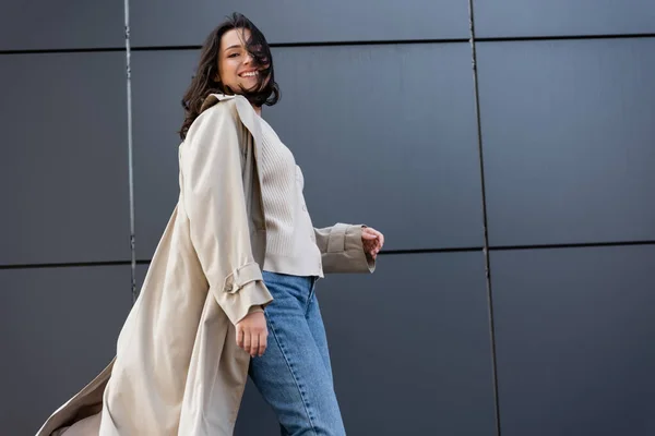 Donna allegra in cappotto beige sorridente alla macchina fotografica mentre in piedi sul vento vicino al muro grigio — Foto stock