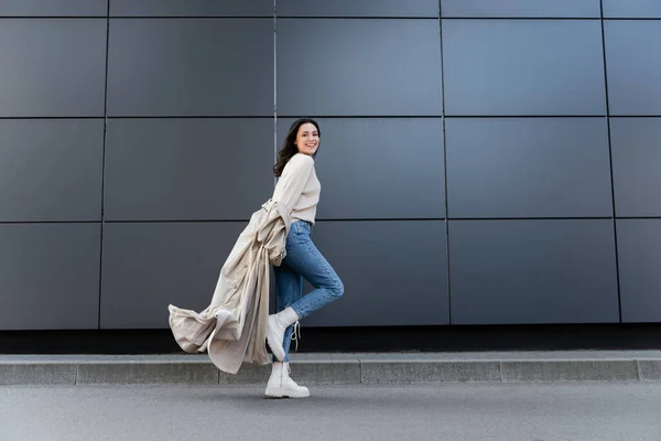 Felice donna in jeans e stivali bianchi sorridente alla fotocamera mentre corre lungo la parete grigia — Foto stock