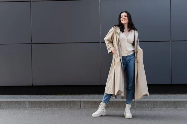 Весела жінка в довгому пальто, джинсах і білих черевиках дивиться далеко біля сірої стіни — стокове фото