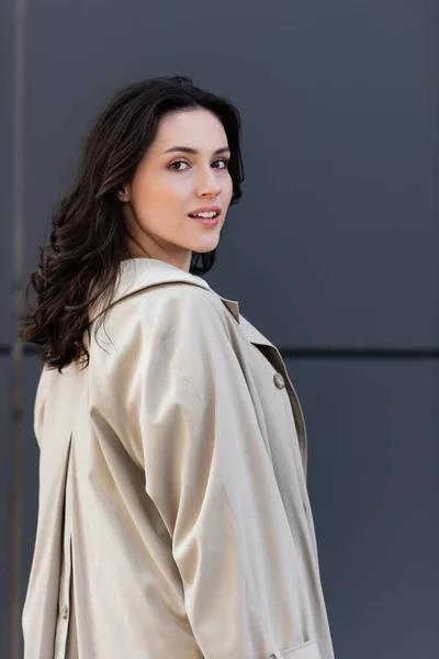 Jeune femme brune en manteau beige regardant la caméra près du mur gris — Photo de stock