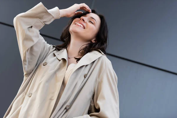Low angle view of smiling woman in beige raincoat posing with closed eyes outdoors — Stock Photo