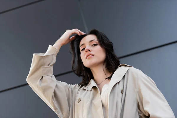 Low angle view of brunette woman in beige coat posing with hand near head outdoors — Stock Photo