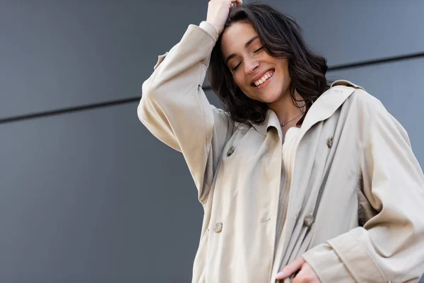 Mulher morena feliz em capa de chuva bege posando com olhos fechados perto da parede cinza — Fotografia de Stock