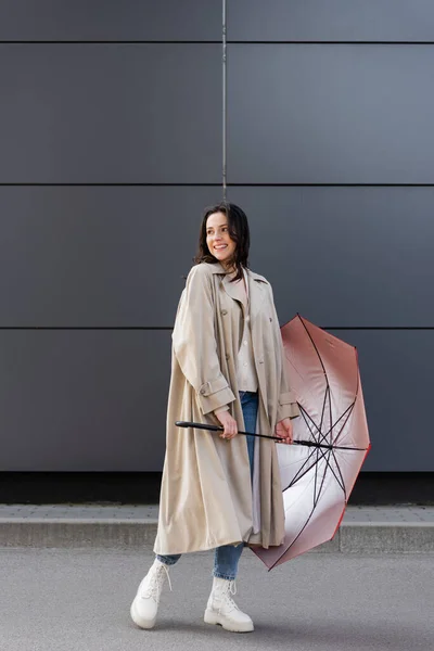 Счастливая женщина в длинном плаще оглядываясь назад, позируя с красным зонтиком — стоковое фото
