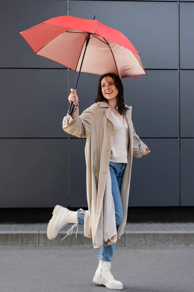 Радісна жінка в стильному осінньому вбранні, стоячи на одній нозі під червоною парасолькою на відкритому повітрі — стокове фото