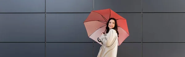 Donna alla moda con ombrello rosso guardando indietro vicino al muro grigio, banner — Foto stock