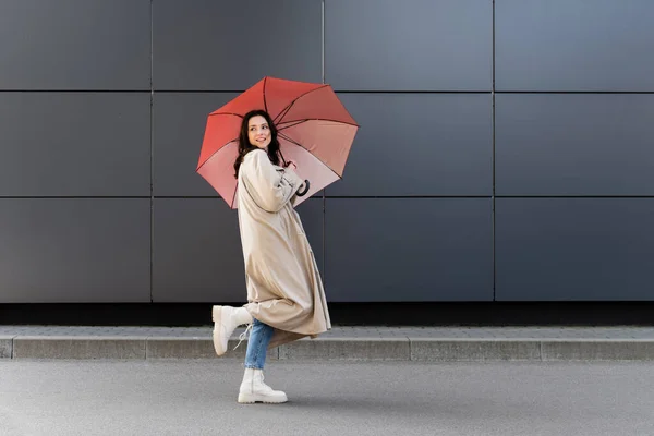 Morena mujer en botas blancas y abrigo beige mirando hacia atrás bajo paraguas rojo - foto de stock