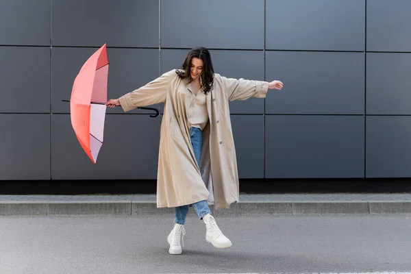 Mulher alegre em capa de chuva longa e botas brancas posando com guarda-chuva vermelho perto da parede cinza — Fotografia de Stock