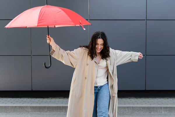 Junge Frau in beigem Regenmantel lächelt, während sie mit rotem Regenschirm in der Nähe der grauen Wand posiert — Stockfoto