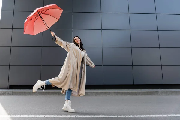 Femme heureuse en manteau beige et bottes blanches posant avec parapluie rouge dans la main levée — Photo de stock