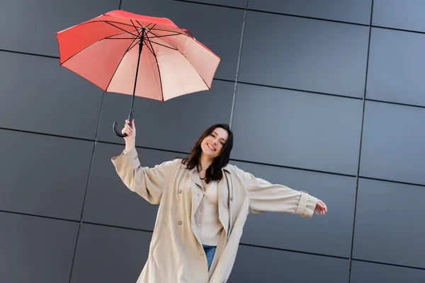 Bruna donna in cappotto beige sorridente alla macchina fotografica sotto l'ombrello rosso vicino al muro grigio — Foto stock