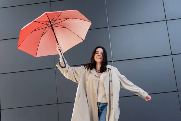 Счастливая брюнетка в бежевом пальто смотрит в камеру, держа красный зонтик рядом с серой стеной — стоковое фото