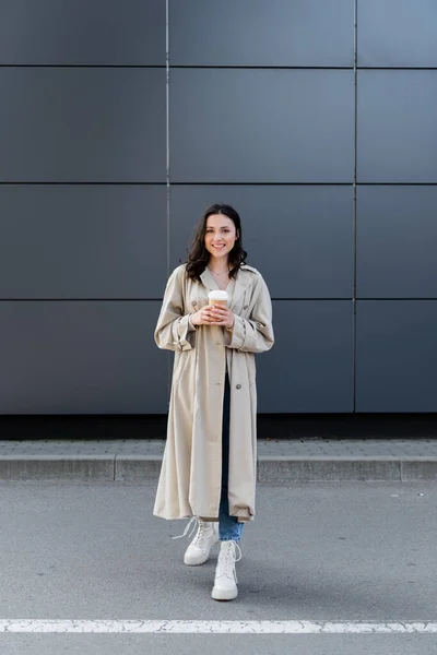 Happy woman in beige raincoat and white boots holding takeaway drink while smiling at camera — Stock Photo