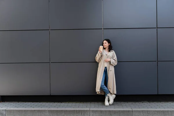 Jovem mulher de capa longa em pé com café para ir perto da parede cinza — Fotografia de Stock