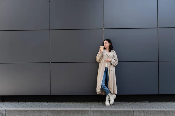 Young woman in long raincoat standing with coffee to go near grey wall — Stock Photo