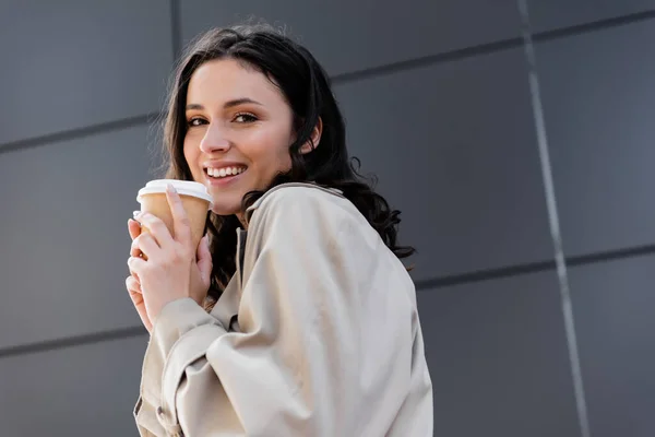 Счастливая женщина с кофе, чтобы пойти улыбаться на камеру возле серой стены за окном — стоковое фото