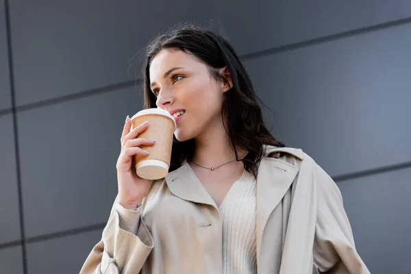 Giovane donna elegante bere caffè per andare mentre guardando lontano vicino muro grigio — Foto stock