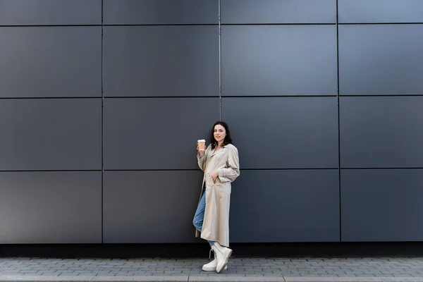 Stylish woman in long coat standing with coffee to go near grey wall — Stock Photo