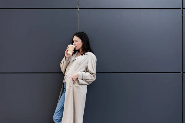 Брюнетка женщина пьет кофе, стоя рядом с серой стеной с рукой в кармане бежевого пальто — стоковое фото