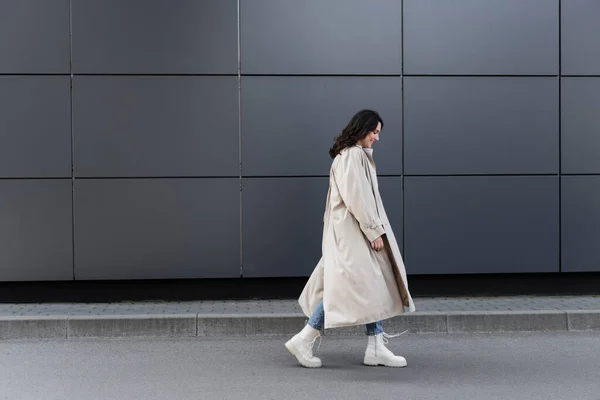 Vista lateral de mujer joven en botas blancas y impermeable largo caminando cerca de la pared gris - foto de stock