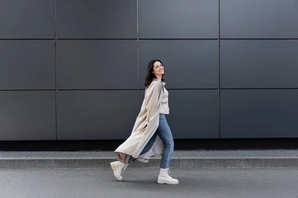 Mujer alegre caminando a lo largo de la pared gris en abrigo largo y botas de cuero blanco - foto de stock