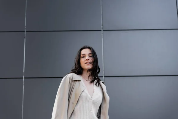 Mujer joven y elegante en impermeable mirando a la cámara cerca de la pared gris - foto de stock