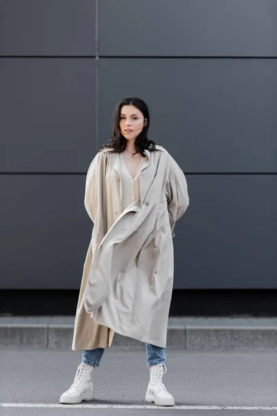 Mulher na moda em casaco bege e botas de couro branco de pé com as mãos atrás das costas — Fotografia de Stock