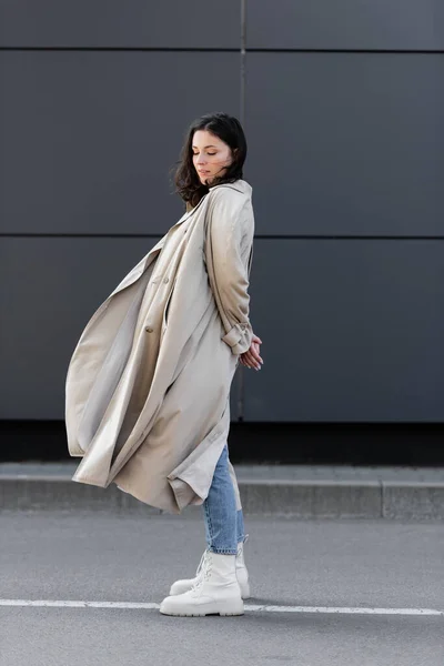 Модна жінка в довгому пальто, що стоїть з закритими очима і руками за сірою стіною — стокове фото