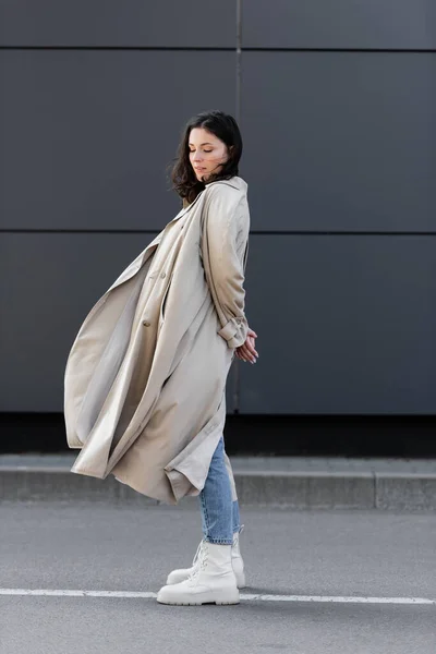 Trendy woman in long coat standing with closed eyes and hands behind back near grey wall — Stock Photo