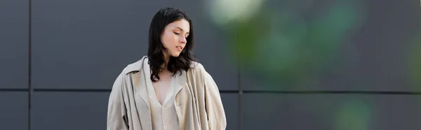 Mujer joven en impermeable beige de pie cerca de la pared gris en primer plano borroso, bandera - foto de stock