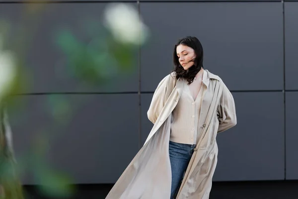 Joven morena mujer en elegante traje de otoño de pie cerca de la pared gris en primer plano borrosa - foto de stock