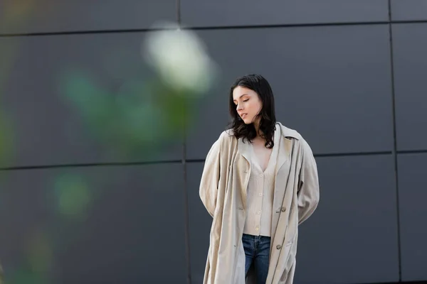 Femme brune en manteau beige posant avec les mains derrière le dos près du mur gris sur le premier plan flou — Photo de stock