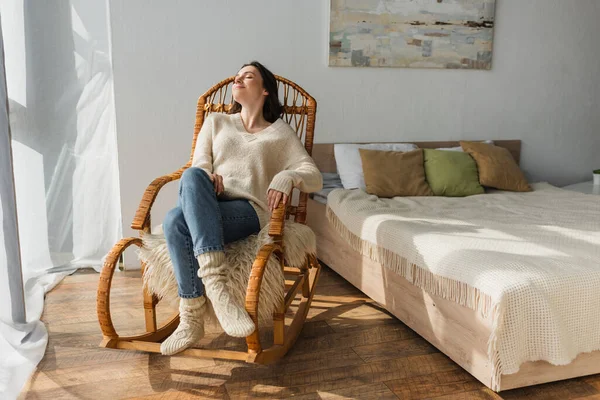 Femme joyeuse en pull doux et chaussettes chaudes reposant dans une chaise berçante dans la chambre — Photo de stock