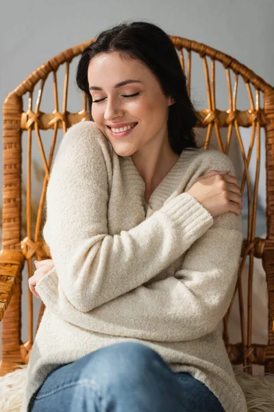 Mujer sonriente en suéter acogedor abrazándose en silla de mimbre - foto de stock