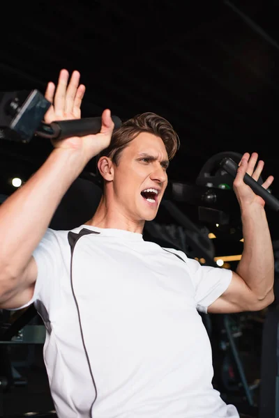 Homem esportivo gritando enquanto treinava na extensão do braço exercitando máquina no centro esportivo — Fotografia de Stock