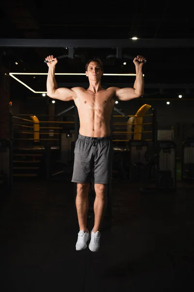 Без рубашки мускулистый мужчина в шортах и кроссовках работает на горизонтальном баре в тренажерном зале — стоковое фото