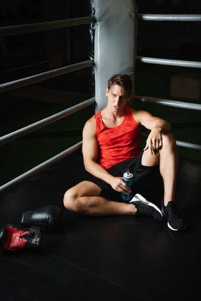Уставший мужчина в спортивной одежде держит спортивную бутылку, сидя на полу рядом с боксерскими перчатками — стоковое фото