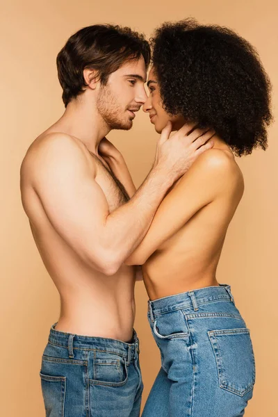 Seitenansicht eines hemdslosen Mannes und einer hispanischen Frau, die sich umarmen, während sie sich isoliert auf beige anlächeln — Stockfoto