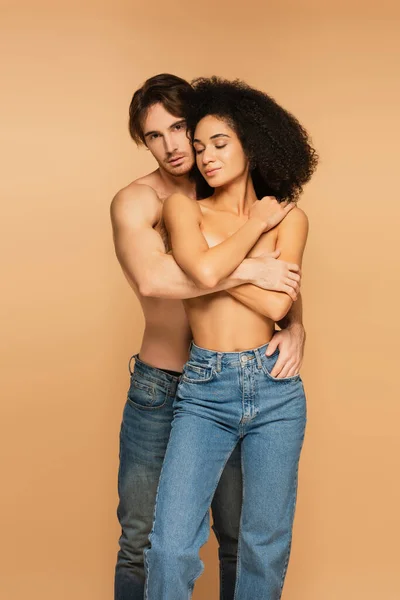 Sexy man embracing topless hispanic woman and holding hand in pocket of her jeans isolated on beige — стоковое фото