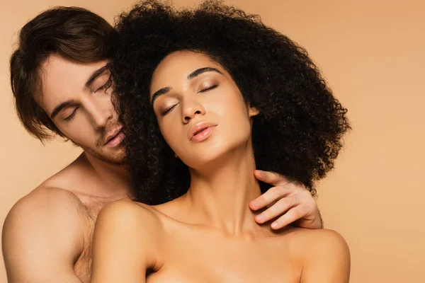 Jeune homme aux yeux fermés embrassant femme latine passionnée et topless isolé sur beige — Photo de stock