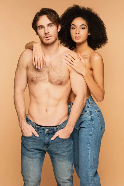 Homem sem camisa de pé com as mãos em bolsos de jeans perto de mulher hispânica sexy abraçando-o isolado no bege — Fotografia de Stock
