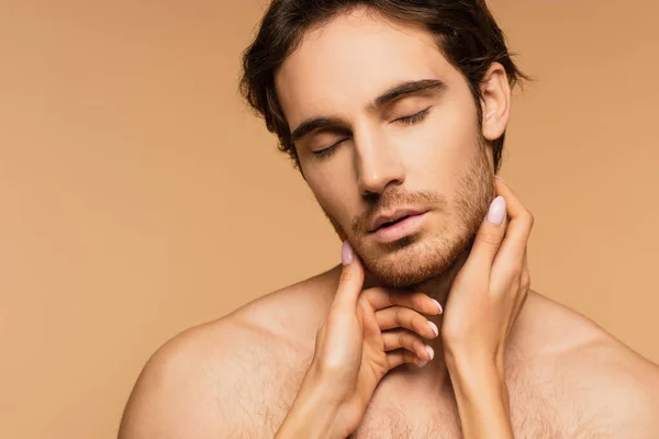 Young shirtless man with closed eyes near female hands touching his face isolated on beige — Stock Photo