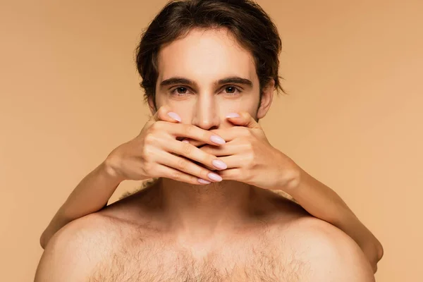 Young shirtless man looking at camera while woman covering his mouth with hands isolated on beige — Stock Photo